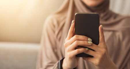 Woman relax at home, chatting. New order and great offer. Smiling muslim woman in hijab typing message on smartphone in living room interior, selective focus on gadget, blurred, cropped, panorama