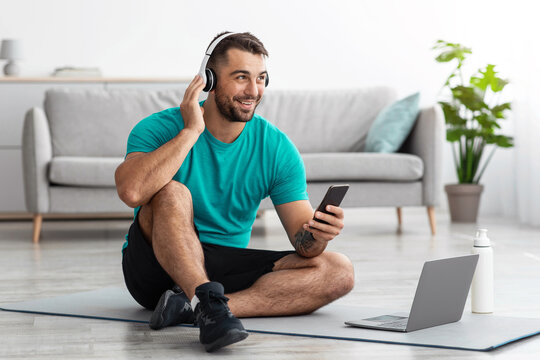 Cheerful millennial caucasian man in headphones sits on mat on floor typing on smartphone, with computer in living room interior. New normal, remote training and technology for sports, video call - Powered by Adobe