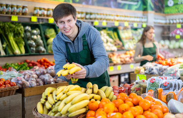 Young salesman in apron lays out bananas on counter in vegetable shop..