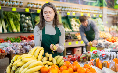 Smiling young saleswoman placing bananas in a market basket in large grocery store