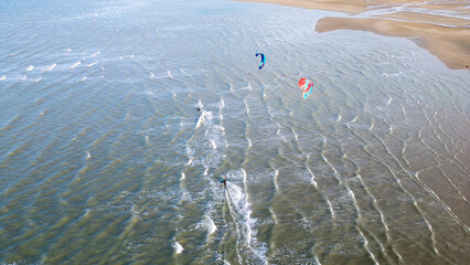 Two kitesurfers with colorful kites speed across the water near the beach. Small waves and steady wind create perfect conditions for watersports, while the aerial perspective highlights both movement