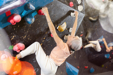 Sporty elderly female exercising in boulder climbing hall without rope, reaching new results, enjoying new challenges in amusement park