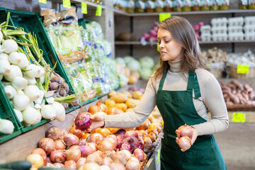 Young woman seller in apron lays out onions on counter in vegetable shop