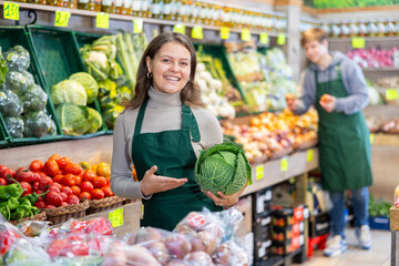 Positive young female seller holding cabbage standing by counter in vegetable market
