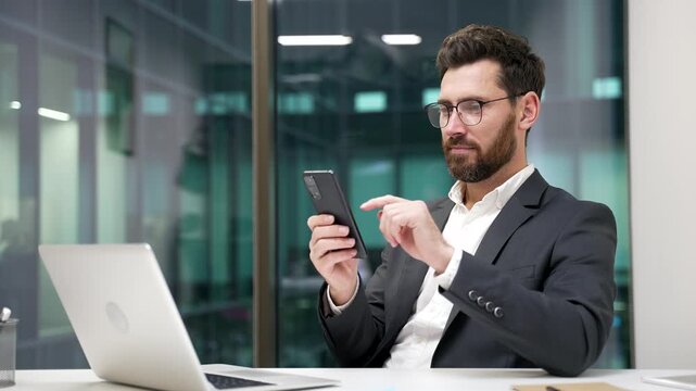 Smiling businessman is using mobile phone while sitting at desk at workplace in modern business office. Happy entrepreneur in formal suit is chatting online or browsing web social media on smartphone