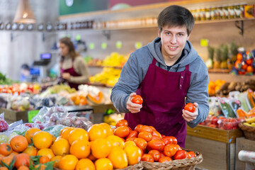 Positive young male seller holding tomatoes standing by counter in vegetable market