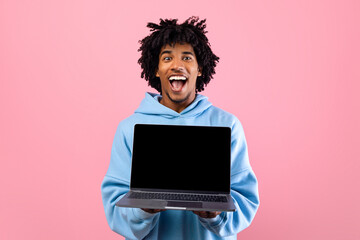 Excited black teen guy holding laptop computer with empty black screen on pink studio background, mockup for website. Cool African American youth demonstrating PC with space for online advertisement