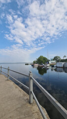 Obraz premium A serene waterfront scene with calm water reflecting the sky. A concrete walkway leads to a dock with boats. Lush greenery is visible nearby.