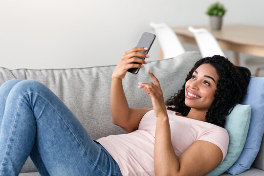 Enjoy spare time alone lying on sofa watching news on phone, comfortable, relaxing at home, using tech. Glad young black woman typing on smartphone, surfing in internet in living room interior - Powered by Adobe
