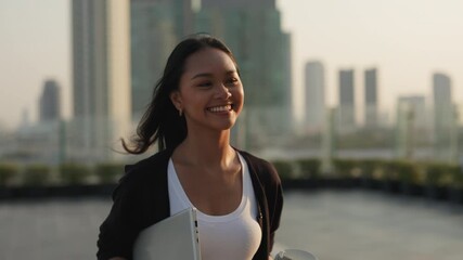 Portrait of joyful Asian businesswoman holds coffee to go and laptop and walks in downtown at sunset. Woman corporate employee with smile expresses professionalism in city setting - Powered by Adobe