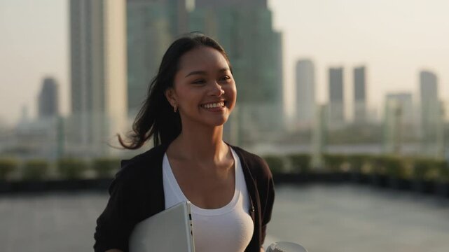 Portrait of joyful Asian businesswoman holds coffee to go and laptop and walks in downtown at sunset. Woman corporate employee with smile expresses professionalism in city setting - Powered by Adobe