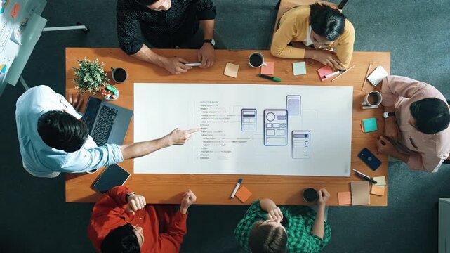 A diverse group of professionals engaged in a brainstorming session around a table, focusing on innovative digital product design and user interface concepts. Raster