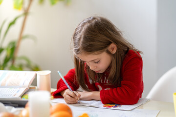 Pretty girl in pajamas sitting at table in kitchen doing homework with focus. She is concentrating while writing, surrounded by study materials, in a cozy home setting
