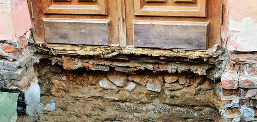 Damaged building foundation with crumbling brick plaster erosion and exposed soil showing decay...