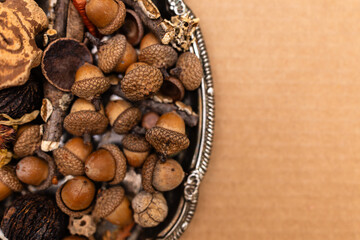 close-up of a collection pile of acorns from an oak tree and various other pieces of nature on a silver platter against a brown background with copy space available in a horizontal orientation  