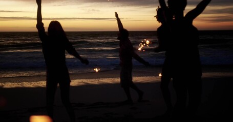 Dancing six friends holding sparklers on wet sandy beach with footprints and ocean waves, at dusk