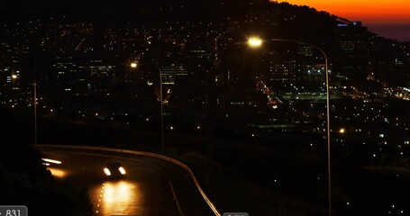 Two cars with headlights navigating wet road at dusk on hillside with guardrail and city lights