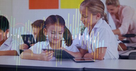 Working two girls wearing polos using tablets in classroom, with smartphone, posters and bar charts