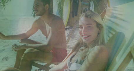Smiling woman in striped bikini sitting on lounge seat at beach palm fronds, surfboards, copy space