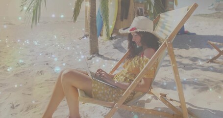 Freelancer wearing straw sun hat and yellow dress working on laptop on sandy beach, with surfboards