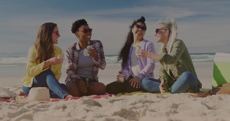 Laughing friends wearing jackets and shades sitting on blanket at beach, sharing snacks and drinks