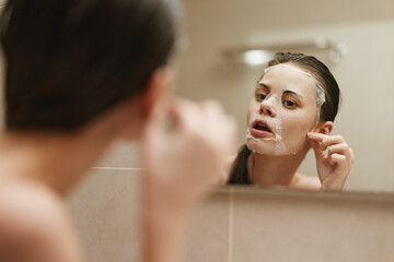 Young woman applying a facial mask in front of a mirror, dedicating time to self-care and wellness....