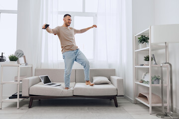 Happy man dancing at home on the sofa, surrounded by bright sunlight. The modern interior creates a joyful and energetic atmosphere for living and leisure.