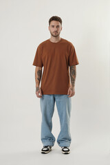 A full-body studio photograph of a young man wearing a rust-colored crew neck t-shirt and light blue baggy jeans. 