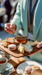 A woman serving a wooden board with cheeses, crackers, macarons, and honey on a sunny day. Selective focus on the honey being drizzled.