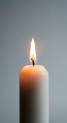 Close-up shot captures the serene ambiance of a lit white candle, its gentle flame casting a warm glow against a soft gray background, evoking feelings of peace and tranquility .