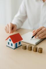 Small house model with red roof and stacks of coins on table with person writing in notebook