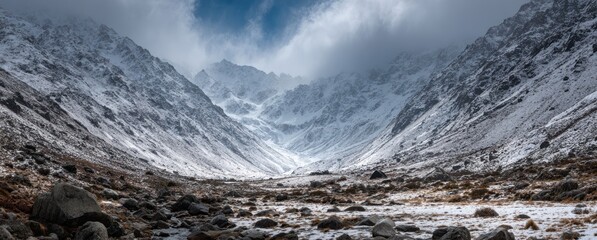 Snow-covered mountains loom in a valley, with rocks scattered on the valley floor and a hint of a stream winding through the landscape
