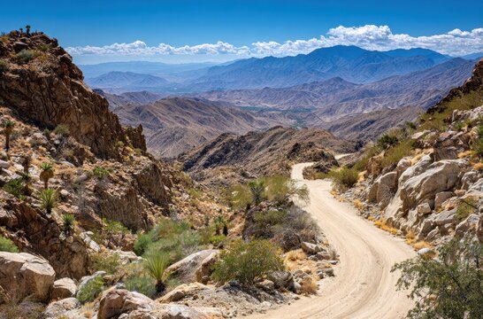 A winding dirt road cuts through rocky hills, revealing a distant mountain range under a bright blue sky and puffy white clouds