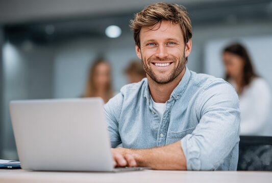 Smiling man in blue shirt works on a laptop at a bright desk with blurred colleagues in the background, conveying a friendly and professional vibe