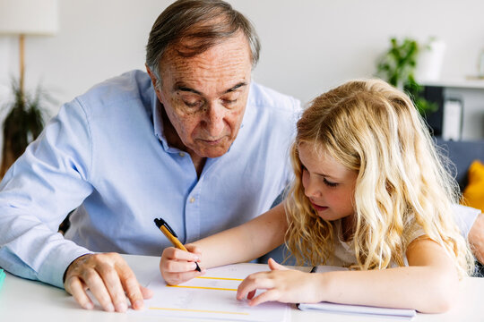 Young little cute girl doing homework with her grandfather at home. Old man helping her daughter with school tasks. Education and family concept. - Powered by Adobe