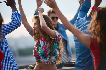 Group of young adults dancing outdoors, multiethnic men and women raising arms and moving energetically, enjoying social gathering with cityscape in background, casual summer setting