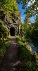 Fototapeta premium A pathway meanders through a lush forest, alongside a stone archway tunnel, on a sunny autumn day.