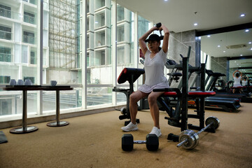 A woman wearing sportswear and a cap is sitting on a gym bench, lifting a dumbbell over her head