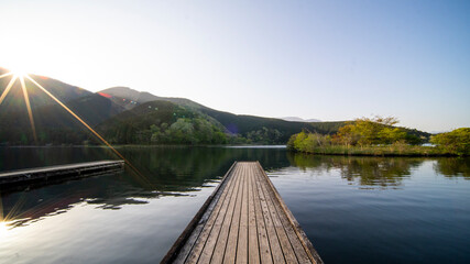 Peaceful wooden pier stretches out over calm lake lush forested hills with the rising sun casting warm glow across the serene morning landscape.