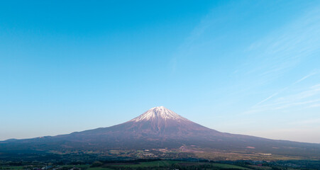 Stunning aerial view of Mount Fuji with its snow-dusted peak towering over a vibrant green forest and wide open fields.