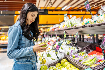 In a bustling shopping mall, Young asian woman joyfully selects fresh fruits and vegetables, examining vibrant options and filling her basket with nutritious choices, selecting care and excitement