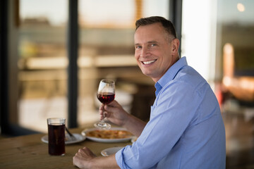Middle-aged man holding red wine glass, smiling at wooden restaurant table with pasta and dark soda