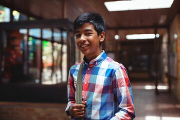 Asian teenage student smiling in school hallway holding shoulder bag strap by artwork, glass cases