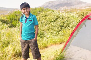 Boy standing next to camping tent with red trim in grassy meadow with shrubs, copy space