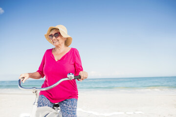 Senior woman standing on beach wearing straw sun hat and sunglasses holding white cruiser bicycle