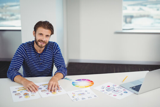 Man arranging color wheel chart and design sheets at modern office desk while using laptop