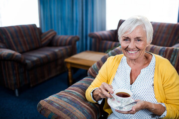 Senior woman holding floral teacup and saucer while sitting in striped armchair at home, copy space