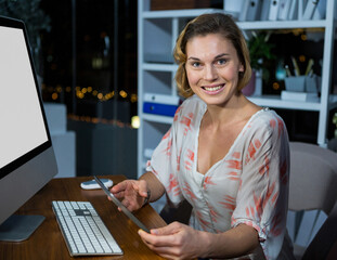 Woman in coral blouse holding tablet and using monitor on wooden desk at night, copy space