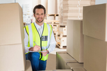 Male warehouse supervisor kneeling among pallets and boxes in storage facility writing on clipboard