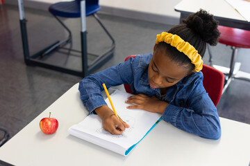 African American child writing in spiral-bound notebook with pencil at school desk near red apple